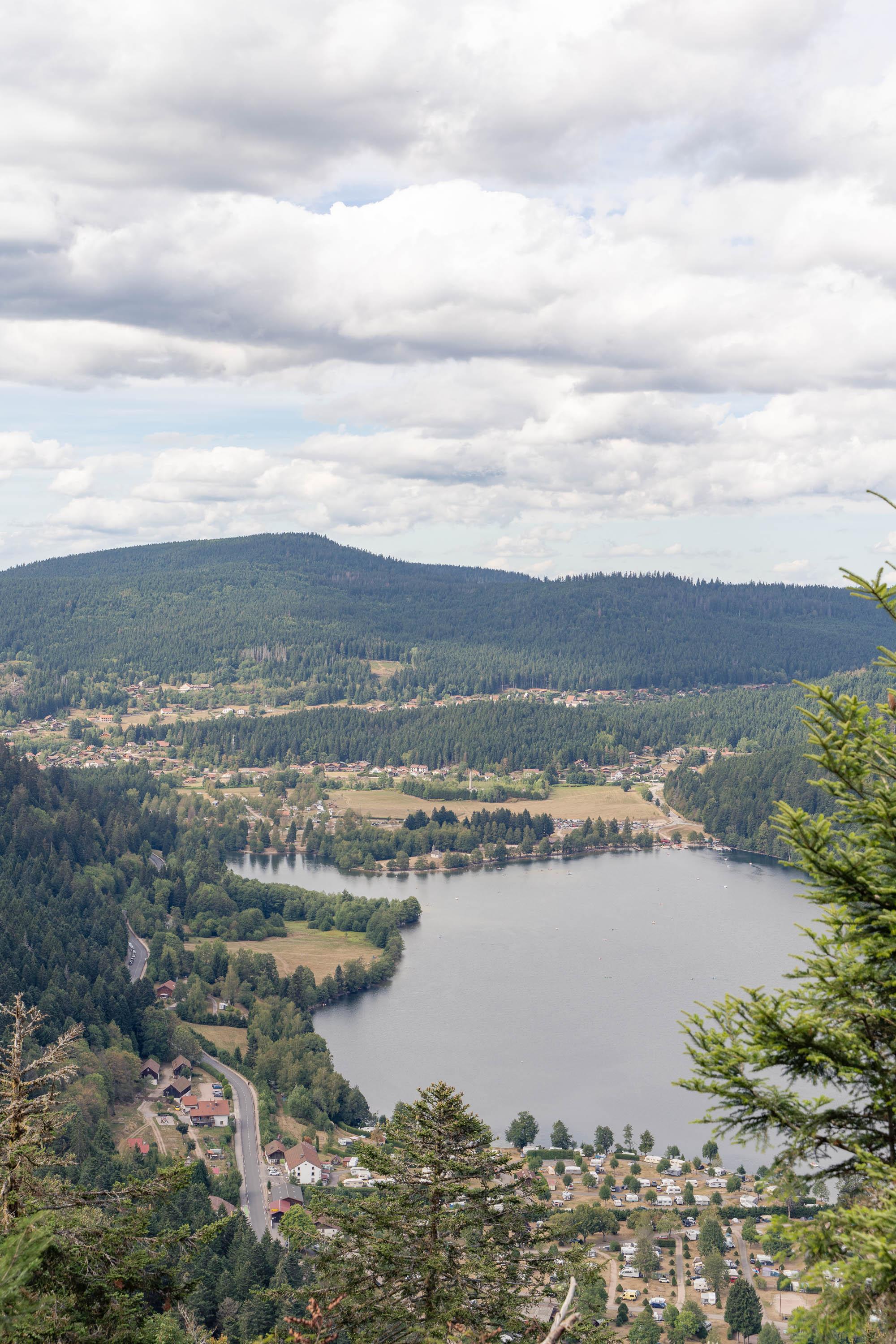 A vertical high-angle photograph shows an expansive valley featuring a lake, surrounding evergreen forests, small settlements, and rolling green hills under an overcast sky.

In the upper third of the image, thick white and light grey clouds fill the sky above a distant, tree-covered mountain ridge that stretches horizontally across the horizon. Below the main ridge, dense green forest covers the surrounding hillsides, interspersed with small clusters of light-colored buildings with red roofs.

In the center and lower half of the frame sits a large, still body of water. The lake's shoreline curves along the left and top edges, meeting grassy fields and patches of forest. Along the lower shore of the lake, a flat area holds a dense arrangement of small white structures, parked camper vans, and small buildings, forming a campsite layout near the water.

To the left of the lake, a paved grey road winds along the base of a forested slope, bordered by scattered red-roofed houses and small r