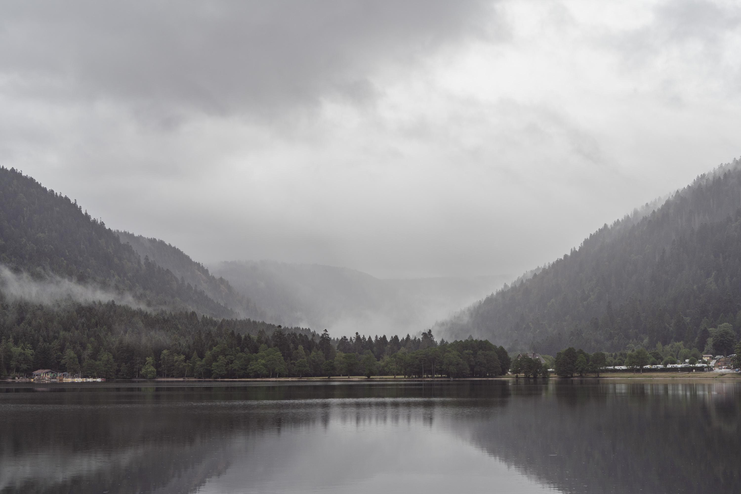 A wide-angle, landscape-oriented photograph depicts a calm, dark lake in the foreground under an overcast, pale gray sky. The dark water reflects the forested mountains and dense green treeline that span the middle of the frame. A low layer of white mist and fog drifts through the tree canopy and along the base of the slopes. Two large, pine-covered mountain slopes rise on the left and right sides, framing a central valley where more distant ridges fade into heavy, low-hanging clouds. Small structures and parked vehicles are visible along the shoreline near the far left and right edges.